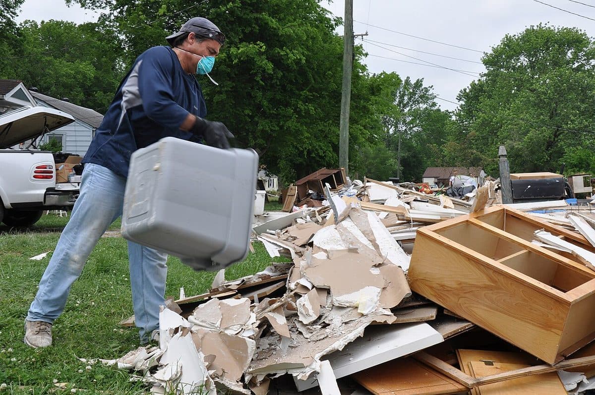 Worker in gloves and dust mask hauling water-damaged drywall and ruined contents from a home to a pickup truck